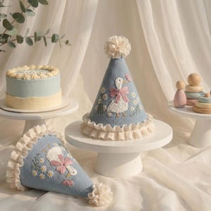 Puede incluir: Dos gorros de fiesta c&oacute;nicos de color azul claro con patos blancos bordados con lazos rosas, rodeados de peque&ntilde;as flores amarillas. Cada gorro est&aacute; rematado con un pomp&oacute;n color crema y un borde de tela con volantes color crema.