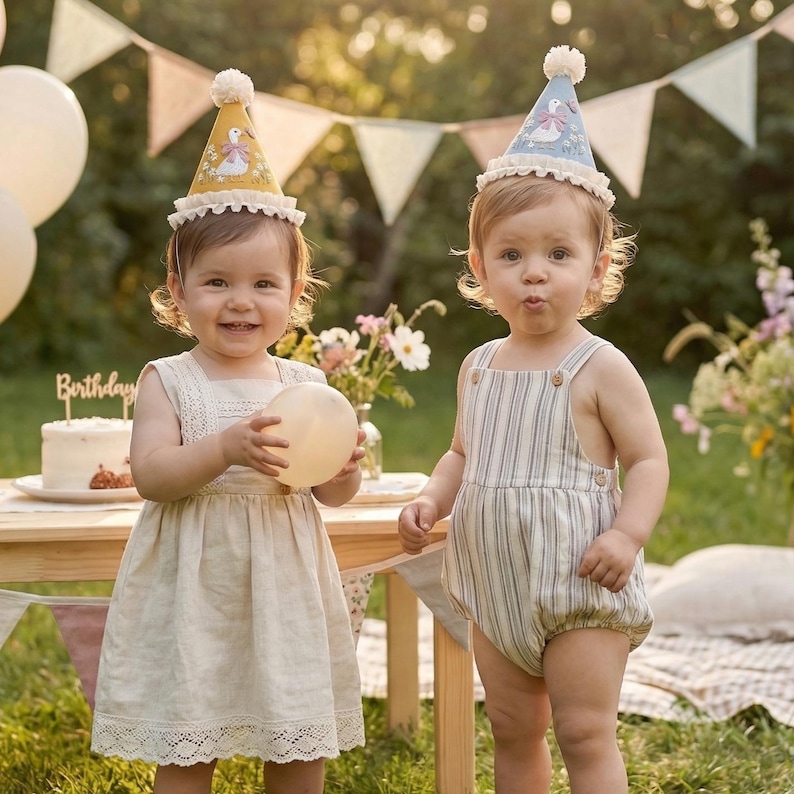Puede incluir: Dos ni&ntilde;os con sombreros de fiesta c&oacute;nicos con una ilustraci&oacute;n de ganso y borde con volantes. Un ni&ntilde;o lleva un vestido beige sin mangas con detalles de encaje, y el otro un pelele a rayas con botones. Un pastel de cumplea&ntilde;os est&aacute; en una mesa.