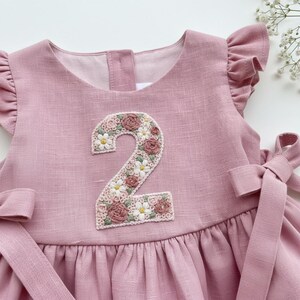 May include: A dusty rose-colored dress with a floral embroidered number 2 on the front. The number is decorated with white daisies and pink roses. The dress has ruffled sleeves and a bow on the side.