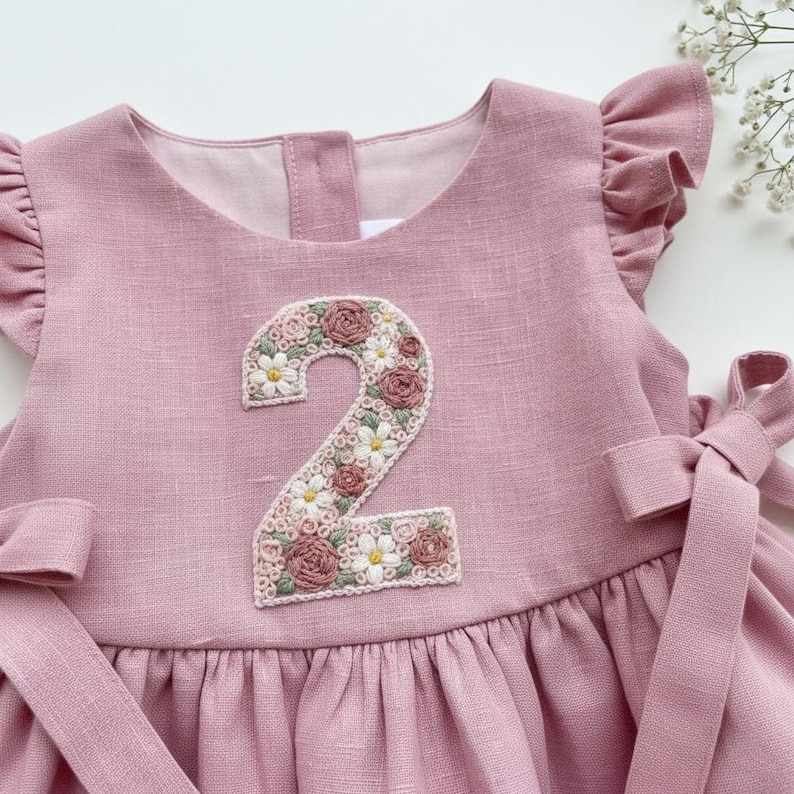 May include: A dusty rose-colored dress with a floral embroidered number 2 on the front. The number is decorated with white daisies and pink roses. The dress has ruffled sleeves and a bow on the side.