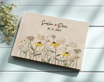 May include: A beige wedding guest book with embroidered floral design. The cover features yellow flowers and greenery, with the names "Suelen & Davi" and the date "16.11.2025" embroidered in dark brown thread. The book is on a light blue wooden surface.