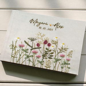 Op de afbeelding: Een lichtgekleurde, rechthoekige doos met geborduurde wilde bloemen. De borduursels bevatten madeliefjes, klavers en ander groen. De namen "Mayara" en "Alex" staan in gouden letters, samen met de datum "10.05.2025".