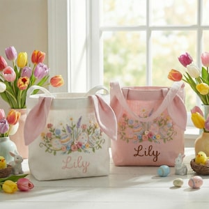 May include: Two Easter-themed tote bags. One is off-white with pink bunny ears and floral embroidery, the other is pink with floral embroidery. Both bags have the name "Lily" embroidered on them. Tulips and Easter decorations are in the background.