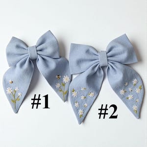 May include: Two light blue fabric hair bows with embroidered floral and bee details. Each bow features a large bow shape with long, flowing tails. The bows are decorated with white daisies and a small bee, with the numbers "#1" and "#2" printed below.