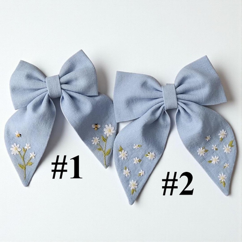 May include: Two light blue fabric hair bows with embroidered floral and bee details. Each bow features a large bow shape with long, flowing tails. The bows are decorated with white daisies and a small bee, with the numbers "#1" and "#2" printed below.