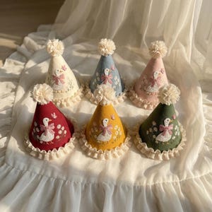 Puede incluir: Seis gorros de fiesta c&oacute;nicos en crema, azul, rosa, rojo, amarillo y verde. Cada gorro presenta un ganso blanco bordado con un lazo rosa, rodeado de margaritas y una mariposa, rematado con un pomp&oacute;n crema y una base de encaje con volantes.