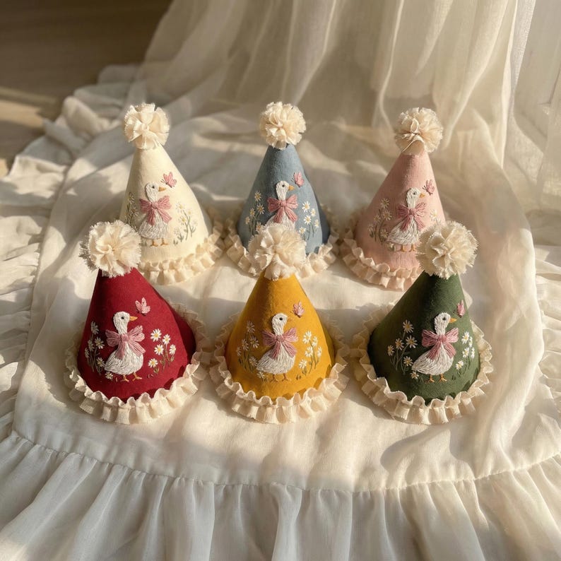 Puede incluir: Seis gorros de fiesta c&oacute;nicos en crema, azul, rosa, rojo, amarillo y verde. Cada gorro presenta un ganso blanco bordado con un lazo rosa, rodeado de margaritas y una mariposa, rematado con un pomp&oacute;n crema y una base de encaje con volantes.