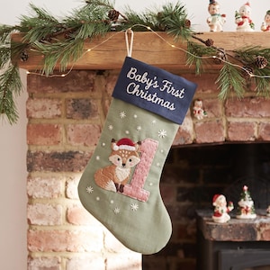 Puede incluir: Un calcetín navideño verde salvia con un puño azul marino con el texto bordado Baby's First Christmas. El calcetín muestra un zorro bordado con un gorro de Papá Noel rojo junto a un número uno grande de color rosa y copos de nieve blancos.