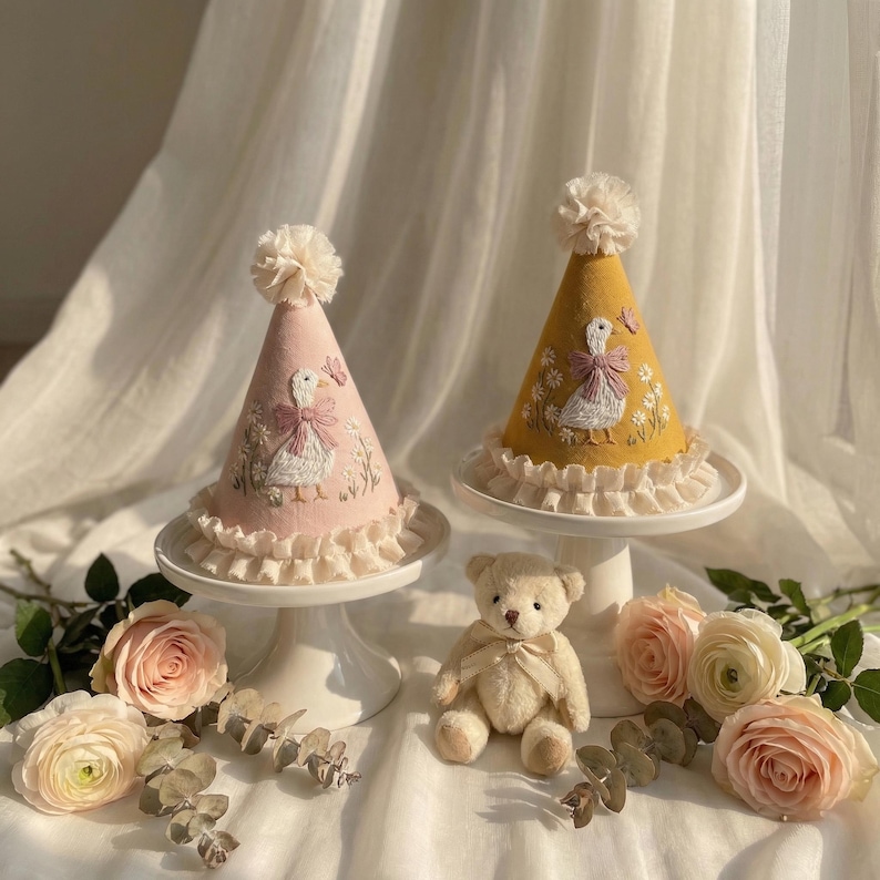 Puede incluir: Dos gorros de fiesta c&oacute;nicos, uno rosa claro y otro amarillo mostaza, cada uno con un pato blanco bordado con un lazo rosa, una peque&ntilde;a mariposa y detalles florales. Ambos gorros tienen un pomp&oacute;n color crema y un borde con volantes.