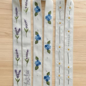 May include: Three white embroidered ribbons with floral designs. One ribbon features lavender, another has blue hydrangeas, and the third showcases daisies. The lavender ribbon has the initials "MG FG" embroidered on it.