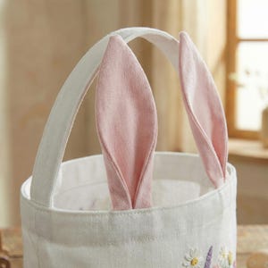 May include: A white fabric Easter basket with a handle and pink bunny ears. The basket has a floral embroidery design around the base. The handle is white and the ears are a soft pink color.