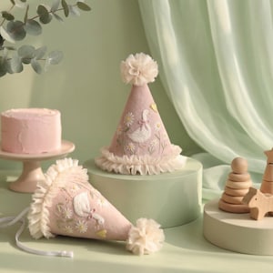 May include: Two conical party hats in dusty pink fabric, each featuring embroidered white geese and yellow daisies. The hats are finished with a ruffled cream-coloured trim at the base and a matching pom-pom at the top, with white ribbon ties.