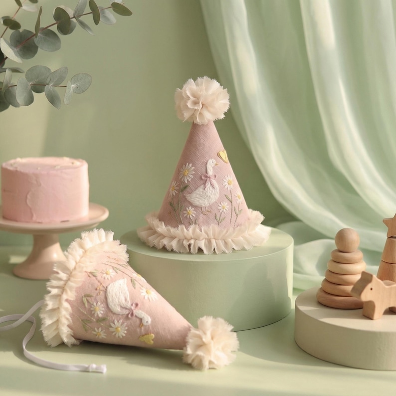 May include: Two conical party hats in dusty pink fabric, each featuring embroidered white geese and yellow daisies. The hats are finished with a ruffled cream-coloured trim at the base and a matching pom-pom at the top, with white ribbon ties.