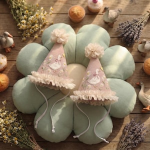 May include: Two cone-shaped party hats in dusty pink fabric, each featuring embroidered white geese, yellow flowers, and green stems. The hats are topped with cream-coloured tulle pom-poms, finished with ruffled tulle trim at the base, and include thin white chin straps.