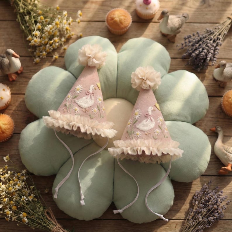 May include: Two cone-shaped party hats in dusty pink fabric, each featuring embroidered white geese, yellow flowers, and green stems. The hats are topped with cream-coloured tulle pom-poms, finished with ruffled tulle trim at the base, and include thin white chin straps.