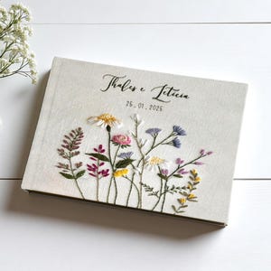 May include: A beige-colored book with embroidered wildflowers in various colors. The cover features the names "Thales e Leticia" and the date "25.01.2025" in script. The book is on a white wooden surface.