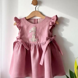 May include: A dusty rose-colored baby dress with a floral embroidered number 1 on the bodice. The dress features ruffled sleeves, a gathered skirt, and bow ties at the waist. It hangs on a wooden hanger.