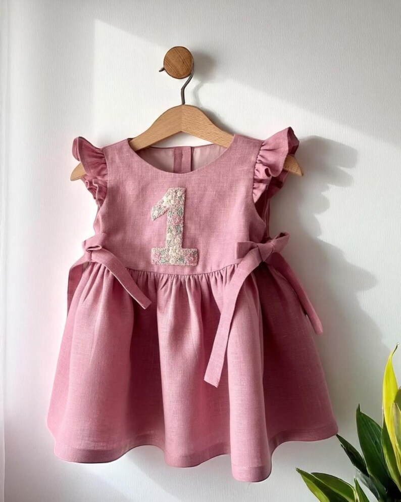 May include: A dusty rose-colored baby dress with a floral embroidered number 1 on the bodice. The dress features ruffled sleeves, a gathered skirt, and bow ties at the waist. It hangs on a wooden hanger.