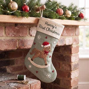 Op de afbeelding: Een saliegroene kerstkous met een geborduurd bruin hobbelpaard dat een rode kerstmuts en een rode strik draagt. De witte boord is geborduurd met de tekst Henry's First Christmas. Witte sneeuwvlokken en rode bessen sieren de stof van de kous.