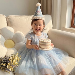 Puede incluir: Un vestido azul claro con corpi&ntilde;o de encaje y falda de tul en capas. El vestido se combina con un gorro de fiesta c&oacute;nico a juego que presenta un pomp&oacute;n blanco, un borde con volantes y un dise&ntilde;o de cisne bordado con peque&ntilde;os detalles florales.