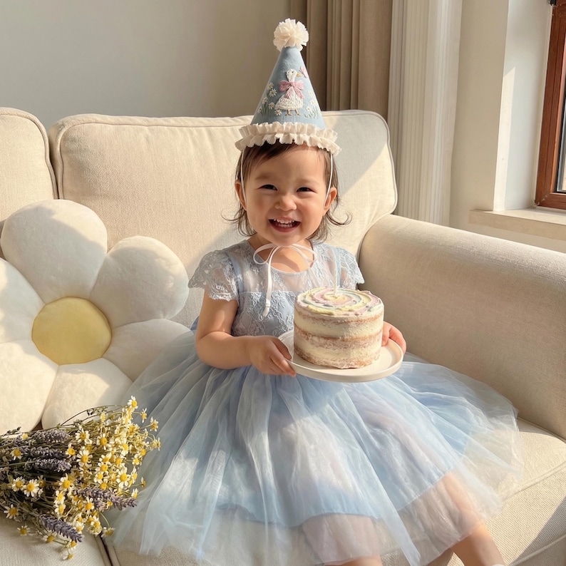 Puede incluir: Un vestido azul claro con corpi&ntilde;o de encaje y falda de tul en capas. El vestido se combina con un gorro de fiesta c&oacute;nico a juego que presenta un pomp&oacute;n blanco, un borde con volantes y un dise&ntilde;o de cisne bordado con peque&ntilde;os detalles florales.