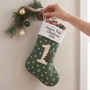 以下が含まれることがあります： Anna's First Christmas 2026という刺繍が施された白いカフ付きの緑色のクリスマスストッキング。ストッキングには、刺繍された緑色のクリスマスツリーと白い雪の結晶模様があしらわれた、大きなクリーム色の数字の1のアップリケが付いています。