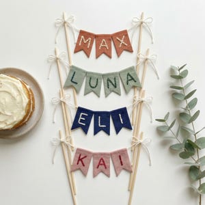May include: Four colorful fabric banner cake toppers with names embroidered on them. The banners are in shades of rust, sage green, navy blue, and dusty rose. Each banner is attached to wooden dowels with white string and bows. A cake is visible on the left.