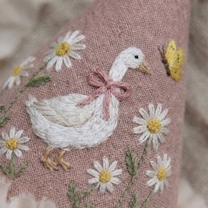 May include: A dusty pink linen fabric featuring detailed embroidery of a white goose wearing a small pink bow around its neck. The scene includes several white daisies with yellow centres and a single yellow butterfly embroidered on the surface.