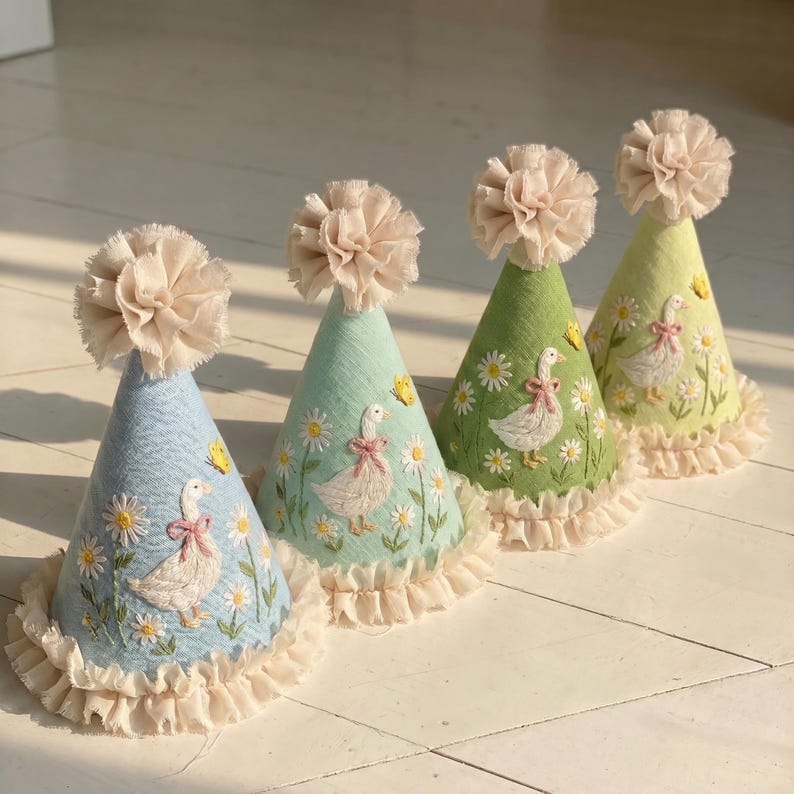 May include: Four cone-shaped party hats in light blue, mint green, sage green, and pale yellow. Each hat features embroidered white geese with pink bows, yellow butterflies, and white daisies. The hats are topped with beige fabric pom-poms.