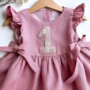 May include: A pink linen dress for a baby or toddler, featuring a floral embroidered number "1" on the bodice. The dress has ruffled sleeves and bows on the shoulders. It is hanging on a wooden hanger.