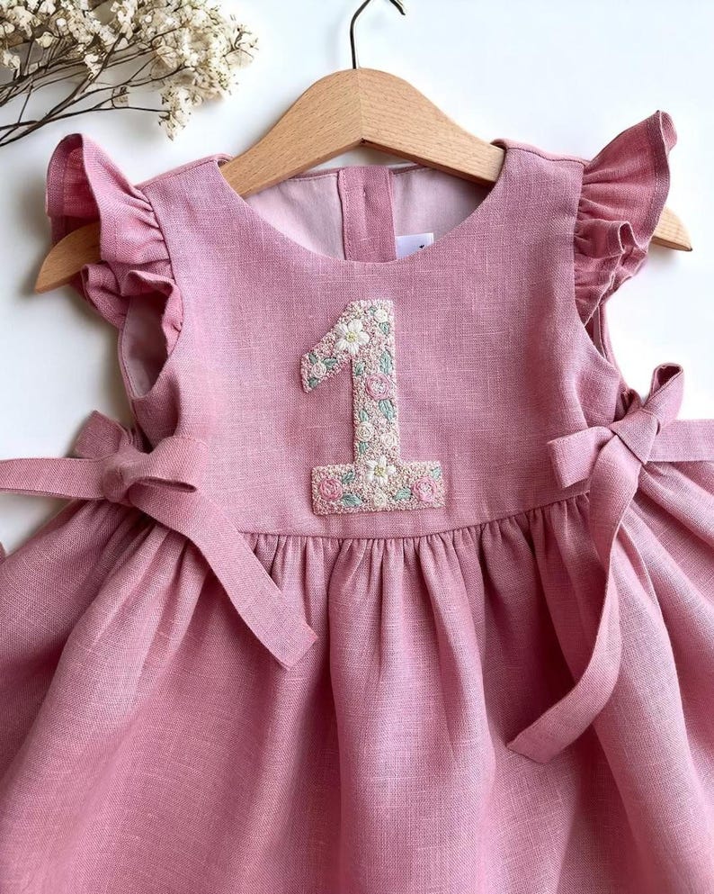 May include: A pink linen dress for a baby or toddler, featuring a floral embroidered number "1" on the bodice. The dress has ruffled sleeves and bows on the shoulders. It is hanging on a wooden hanger.