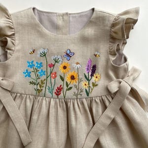 May include: A beige linen dress with embroidered wildflowers, butterflies, and bees. The dress features ruffled sleeves and tie details at the sides. The embroidery includes blue, yellow, pink, and purple flowers.