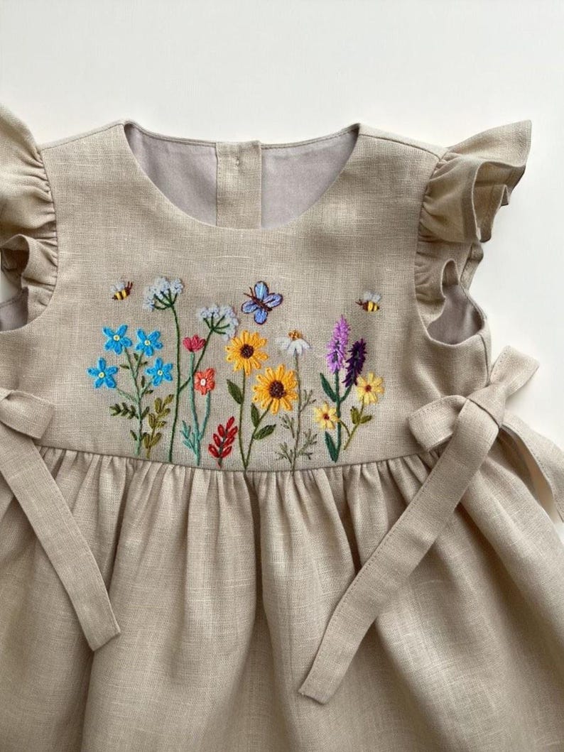 May include: A beige linen dress with embroidered wildflowers, butterflies, and bees. The dress features ruffled sleeves and tie details at the sides. The embroidery includes blue, yellow, pink, and purple flowers.