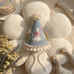 Puede incluir: Un gorro de fiesta c&oacute;nico de color azul claro con margaritas blancas bordadas y una oca blanca central con un lazo rosa. El gorro est&aacute; rematado con un peque&ntilde;o pomp&oacute;n de tela, un borde de volantes color crema en la base y dos finas cintas de tela blanca para atar.