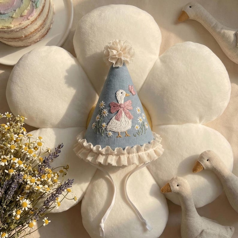 Puede incluir: Un gorro de fiesta c&oacute;nico de color azul claro con margaritas blancas bordadas y una oca blanca central con un lazo rosa. El gorro est&aacute; rematado con un peque&ntilde;o pomp&oacute;n de tela, un borde de volantes color crema en la base y dos finas cintas de tela blanca para atar.