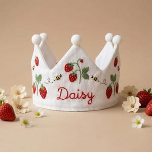 以下が含まれることがあります： 白い布製の王冠で、赤いイチゴ、緑の葉、小さな黄色と黒のハチが刺繍されています。前面には赤い筆記体で「Daisy」という名前が刺繍されています。王冠の先端には小さな白いポンポンが付いています。