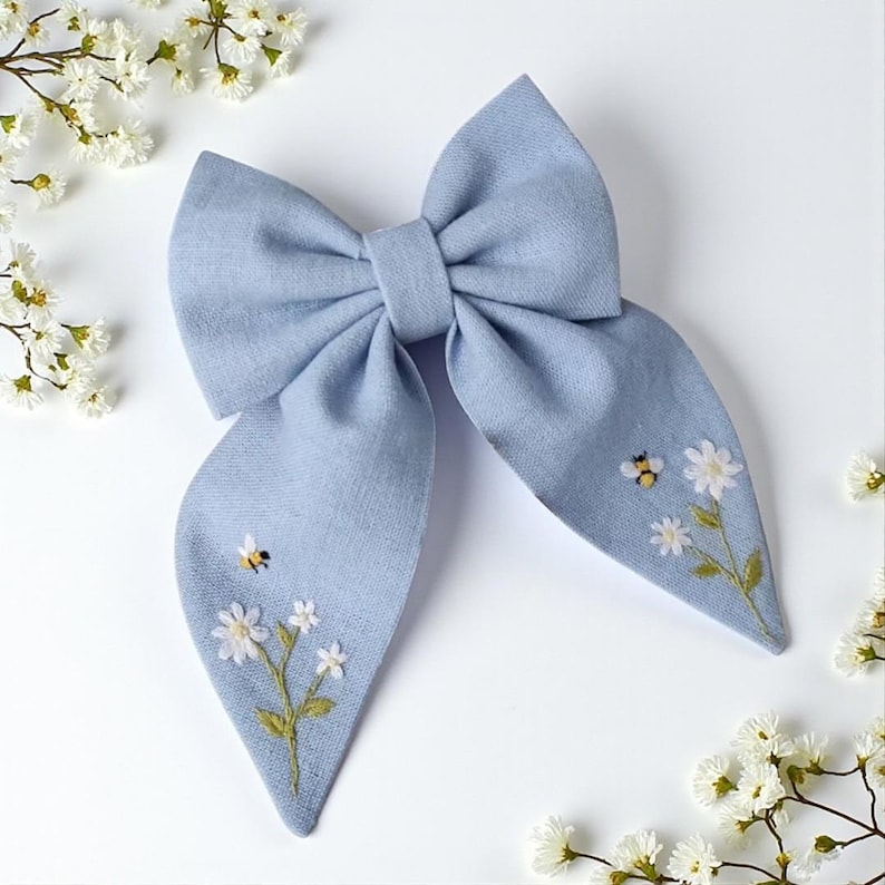 May include: A light blue fabric hair bow with embroidered bees and white flowers. The bow has a classic bow shape with long, flowing tails. The embroidery features yellow and black bees and delicate white flowers with green stems and leaves.