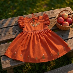 May include: An orange dress with embroidered floral details and ruffled sleeves. The dress is displayed on a wooden bench next to a basket of red apples. The dress has a round neckline and a gathered skirt.