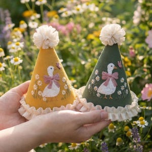Puede incluir: Dos gorros de fiesta c&oacute;nicos, uno amarillo mostaza y otro verde bosque, cada uno con una oca blanca bordada con un lazo rosa, peque&ntilde;as flores, un borde con volantes color crema en la base y un pomp&oacute;n color crema esponjoso en la parte superior.
