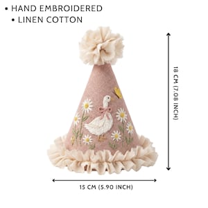 May include: A cone-shaped party hat made of dusty pink linen cotton, featuring hand-embroidered white daisies, a goose with a pink bow, and a yellow butterfly. It has a ruffled base and a top pom-pom. Dimensions are 18 cm high by 15 cm wide.