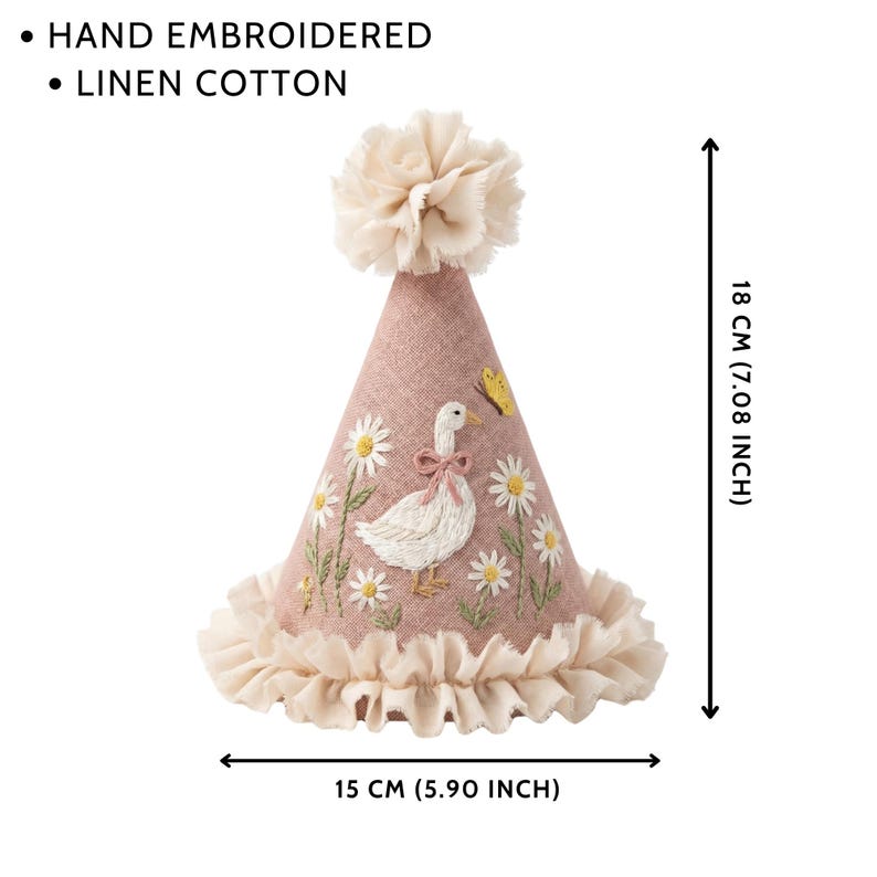 May include: A cone-shaped party hat made of dusty pink linen cotton, featuring hand-embroidered white daisies, a goose with a pink bow, and a yellow butterfly. It has a ruffled base and a top pom-pom. Dimensions are 18 cm high by 15 cm wide.