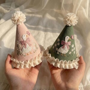 Puede incluir: Dos gorros de fiesta c&oacute;nicos, uno rosa claro y otro verde salvia, con bordados de gansos blancos con lazos rosas, peque&ntilde;as flores y mariposas. Ambos gorros tienen un borde de volantes de color crema y un pomp&oacute;n en la parte superior.