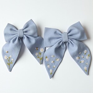 May include: Two light blue fabric hair bows. Each bow features a large bow shape with long, flowing tails. The tails are adorned with embroidered white daisies and a small bee. The bows are set against a white background.