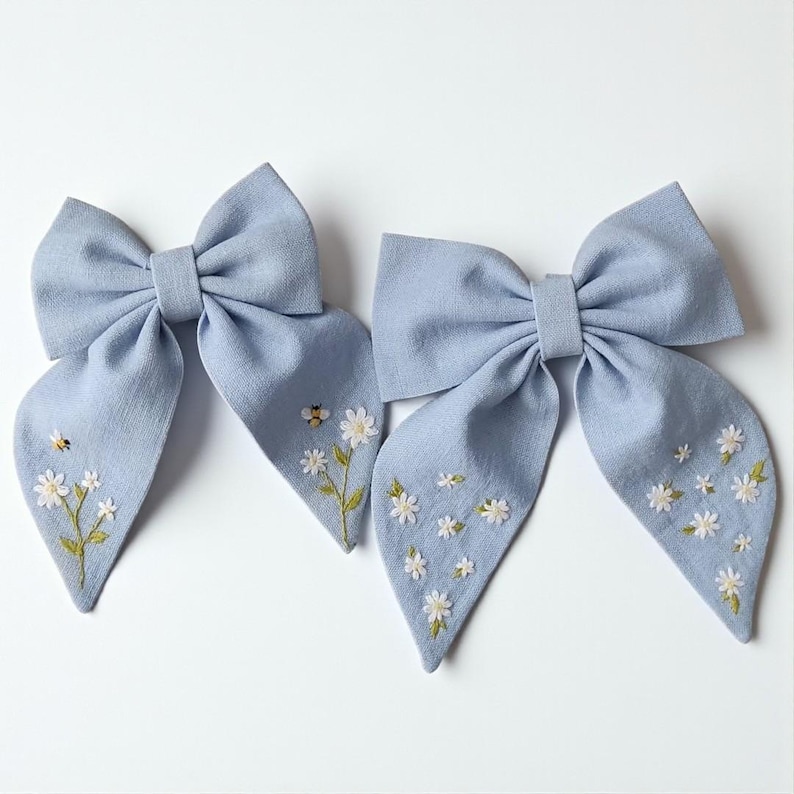 May include: Two light blue fabric hair bows. Each bow features a large bow shape with long, flowing tails. The tails are adorned with embroidered white daisies and a small bee. The bows are set against a white background.