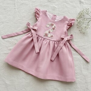 May include: A light pink linen dress for a toddler, featuring a floral embroidered number "2" on the bodice. The dress has flutter sleeves and a bow at the waist. The dress is approximately 24 inches long.
