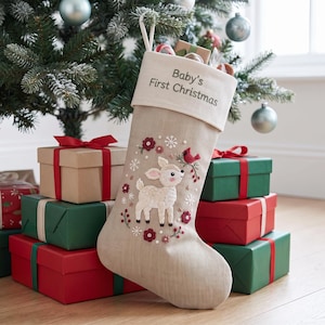 May include: A beige Christmas stocking with a white cuff featuring the text Baby's First Christmas. The stocking is embroidered with a fawn, red flowers, and a cardinal bird. The design includes small white snowflakes and green leaf accents.