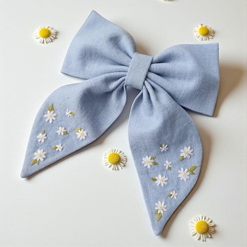 May include: A light blue fabric bow with embroidered white daisies and green leaves. The bow is centered and surrounded by small daisy embellishments. The bow is a hair accessory.