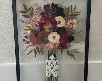 May include: A framed floral arrangement featuring a bouquet of dried flowers in shades of burgundy, cream, and black. The bouquet is wrapped with a green ribbon and an ornate white lace design. The frame is black.