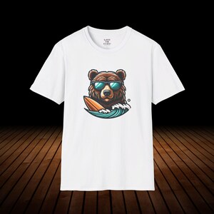 May include: White t-shirt with a graphic of a brown bear wearing sunglasses and riding a surfboard on a wave.