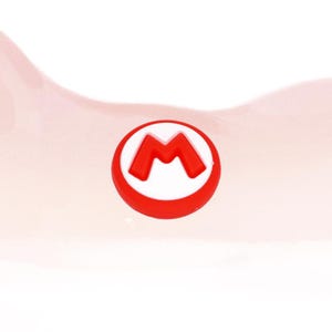 Può includere: Un emblema circolare rosso e bianco con un logo "M" rosso stilizzato. L'emblema è posto su uno sfondo rosa chiaro e morbido, creando un design semplice ma accattivante. Il logo è probabilmente di un videogioco popolare.